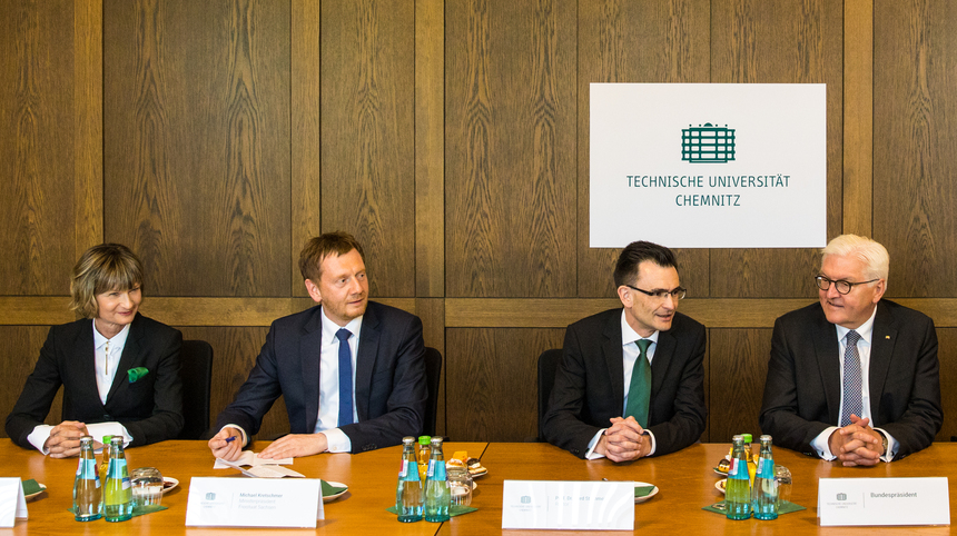 Eine Frau und drei Männer sitzen an einem Tisch. Dahinter sieht man das Logo der Technischen Universität Chemnitz